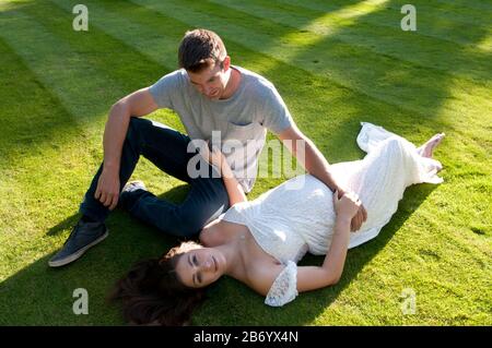 Belle jeune femme enceinte, portant une longue robe blanche, posée sur l'herbe avec son partenaire assis à côté d'elle Banque D'Images