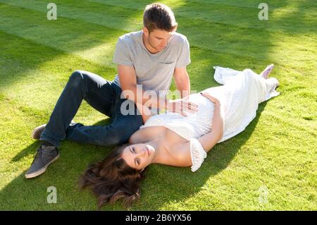 Belle jeune femme enceinte, portant une longue robe blanche, posée sur l'herbe avec son partenaire assis à côté d'elle Banque D'Images