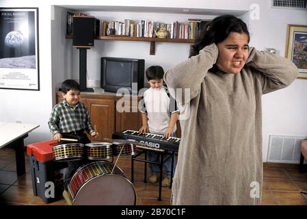 Les jeunes garçons jouent de la musique forte à la maison, tandis que la sœur adolescente couvre ses oreilles pour bloquer le bruit. M. ©Bob Daemmrich Banque D'Images