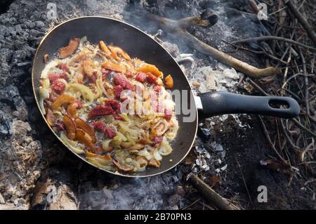 Des tranches de bacon frire dans sa propre graisse avec oignon et saucisse sur le vieux frypan noir. Cuisine en plein air dans la forêt sur feu de camp. Banque D'Images
