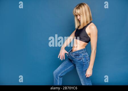 Femme blonde mince dans un large Jean sur fond bleu, espace pour le texte. Perte de poids, fitnes, concept de lipoaspiration. Banque D'Images