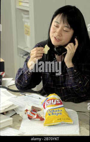 Habitudes alimentaires malsaines. La jeune femme américano-asiatique saute le déjeuner au travail et mange des chips de collation à la place. M. ©Bob Daemmrich Banque D'Images