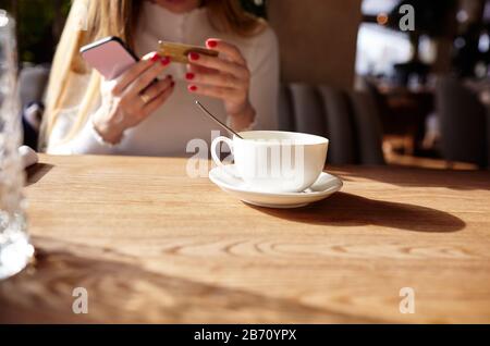 Les mains de la femme tenant le téléphone mobile et la carte de crédit dans le café. Pause café. Tasse de café sur la table. Image floue, mise au point sélective Banque D'Images