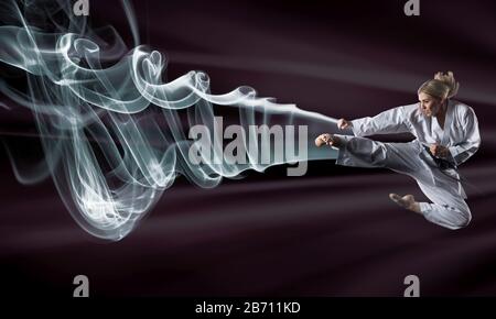 Image composite d'une fille de karaté dans un kimono blanc avec ceinture noire en saut. Fond violet et nuages de fumée Banque D'Images