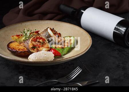 Petits pains de dorado, courgettes et pommes de terre sur une assiette à fond sombre et une bouteille de vin. Banque D'Images