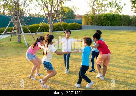 Un grand groupe d'heureux amis asiatiques souriants de jardin d'enfants qui tiennent les mains jouer et danser jouent à la roundelay et se tiennent en cercle dans le parc sur le g Banque D'Images