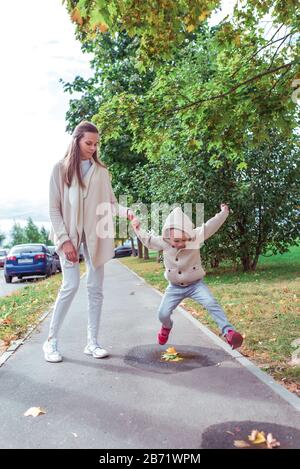 Femme maman marche avec son enfant, en automne en ville, petit garçon sautant à travers des flaques sur le pavé. Reposez-vous pour une promenade le week-end. Vêtements chauds décontractés. Banque D'Images