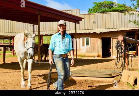 Les chevaux en stabilité sont allés à pied jusqu'au corral par une jeune cow-girl souriante et attrayante à un Ranch Gunstock de 900 acres, île d'Oahu, Laie, Hawaï, États-Unis Banque D'Images