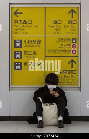 Un jeune homme, portant un masque chirurgical en papier, s'assoit sur le sol pour vérifier son téléphone à la station Shinjuku, Shinjuku, Tokyo, Japon. Banque D'Images