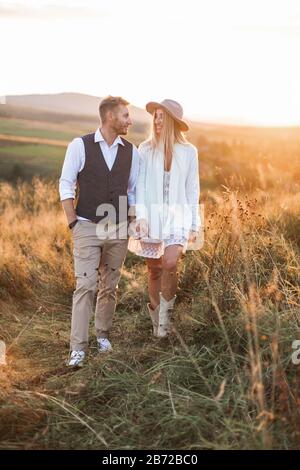 Bel homme élégant dans un costume rustique et jolie femme boho en robe, veste, chapeau et bottes de cowboy, marchant dans le champ, tenant les mains, regardant chacun Banque D'Images