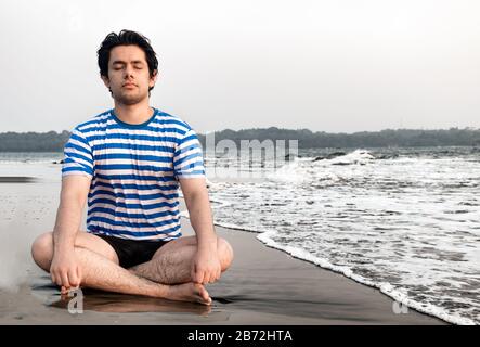 Beau garçon de bonne apparence au début des années 20, médite pour le bien-être physique et mental, tout en étant assis en position lotus sur la plage. Yoga, santé Banque D'Images