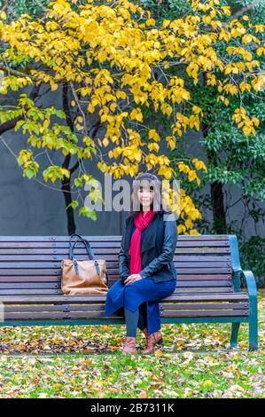 Vue sur le touriste féminin avec écharpe rouge et sac à main en cuir assis sur un banc de parc sous des arbres d'automne à Fitzroy Park, Melbourne, Victoria. Banque D'Images