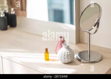 Coiffeuse avec miroir circulaire, petit vase fleuri dans un vase en céramique brillant blanc et rose et flacon de réactif sur le comptoir en bois. Banque D'Images