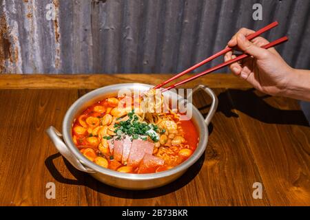 Nouilles à pincer à la main avec baguettes. Soupe traditionnelle coréenne de ramen avec kimchi, jambon, saucisse et fromage dans un bol en argent sur table en bois. Banque D'Images