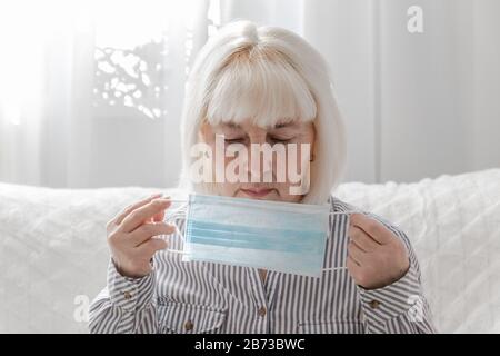 Femme blonde adulte dans une chemise rayée place un masque médical sur son visage dans une pièce à la maison. Protection antivirus, coronavirus, concept ARVI Banque D'Images