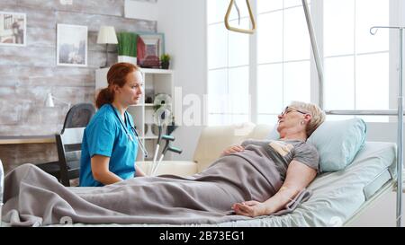 Infirmière dans la maison de retraite parler avec une vieille dame posée dans le lit d'hôpital. De grandes fenêtres avec lumière vive sont derrière Banque D'Images