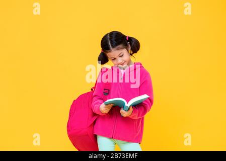 Portrait d'une jolie fille avec des cheveux en queue de porc dans une veste rose et un livre de lecture sac à dos dans un fond de studio isolé jaune Banque D'Images