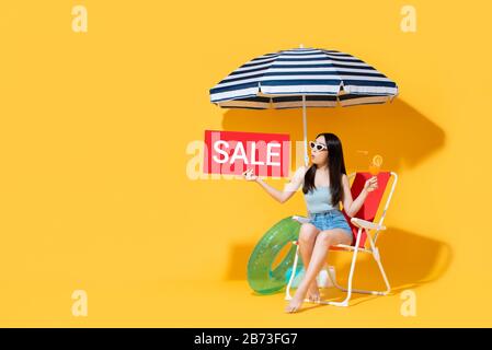 Surprise belle femme asiatique en été tenue tenant le signe de vente rouge tout en étant assis sur chaise de plage isolée sur fond jaune avec espace de copie Banque D'Images
