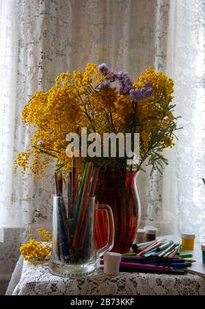 Crayons de couleur, marqueurs et peintures à côté d'un bouquet de Mimosa dans un vase rouge sur un tablecloth en dentelle . Banque D'Images