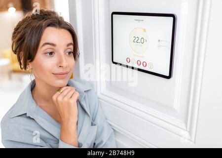 Jeune femme contrôlant la température dans le salon avec un écran tactile numérique. Concept de contrôle du chauffage dans une maison intelligente Banque D'Images