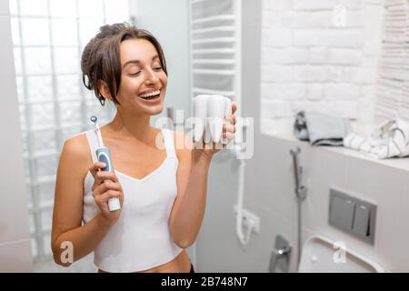 Portrait d'une jeune et gaie femme debout avec brosse à dents électrique pendant les procédures d'hygiène matinales dans la salle de bains Banque D'Images