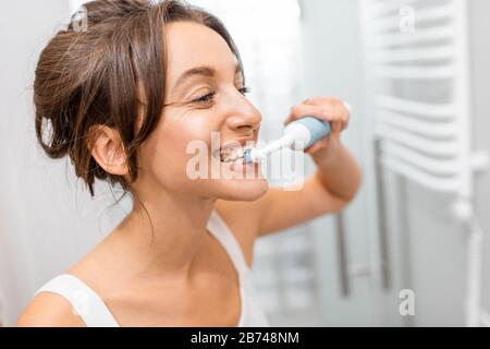 Jeune et gaie femme se brossant les dents avec brosse à dents électrique pendant les procédures d'hygiène matinale dans la salle de bains, portrait du visage Banque D'Images