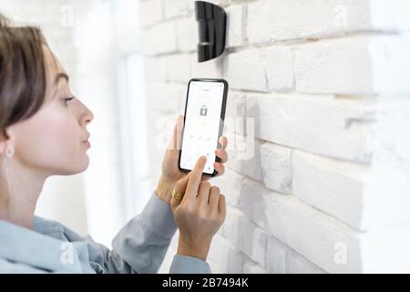 Femme contrôlant le système d'alarme avec smartphone et application mobile spéciale sans fil, debout près du capteur de mouvement à l'intérieur Banque D'Images