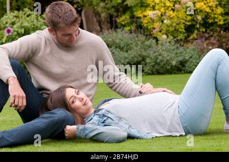 Belle jeune femme enceinte posée sur l'herbe avec son partenaire assis à côté d'elle Banque D'Images