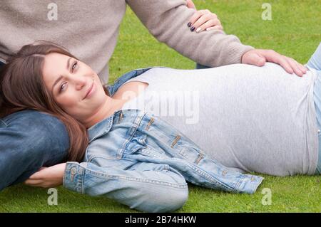 Gros plan, belle jeune femme enceinte posée sur l'herbe avec son partenaire assis à côté d'elle Banque D'Images