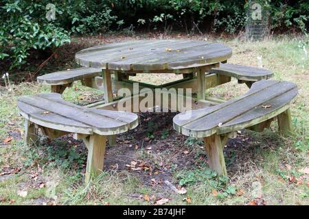 Table ronde de pique-nique en bois avec sièges arrondis sur l'herbe avec sol en dessous et arbustes en arrière-plan. Banque D'Images