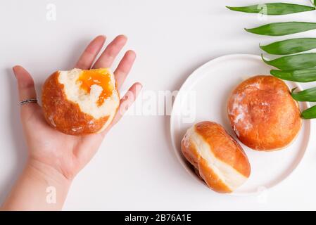 La moitié d'un délicieux beignet de sucre maison fraîchement cuit avec de la confiture d'abricot dans une main de femme et sur une plaque avec des feuilles de palmier au-dessus du fond de papier blanc Banque D'Images