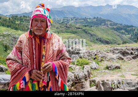 Cusco, Pérou; 24 janvier 2017: L'homme ancien Quechua vêtu d'un poncho coloré et d'un chapeau de Chullo, joue sur l'instrument de musique quena avec une vue de t Banque D'Images