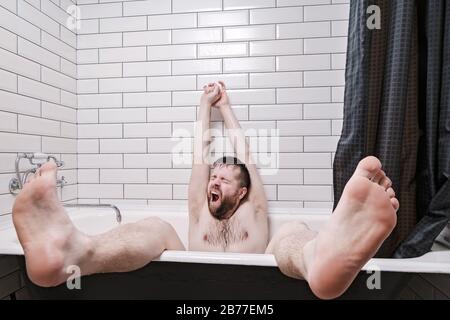L'homme se tient relaxement dans un bain en fonte avec des jambes pendantes, des yawns et s'étire, après les procédures d'eau. Style de vie. Banque D'Images