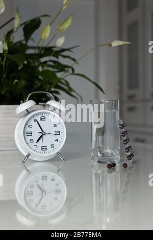 Réveil blanc et pilules médicales avec un verre d'eau sur la table. Santé et médecine. Vitamines et compléments minéraux en capsules. Banque D'Images