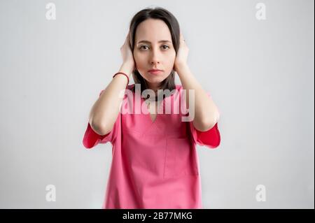 Jeune femme médecin infirmière ou médecin vêtu de vêtements d'hôpital roses, avec des cheveux bruns, dans l'entendre no Evil, posant sur un blanc isolé Banque D'Images