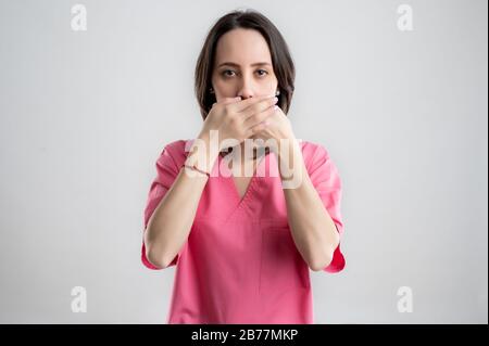 Jeune femme médecin infirmière ou médecin vêtu de vêtements d'hôpital roses, avec des cheveux bruns, dans la parole no Evil posant sur un blanc isolé Banque D'Images