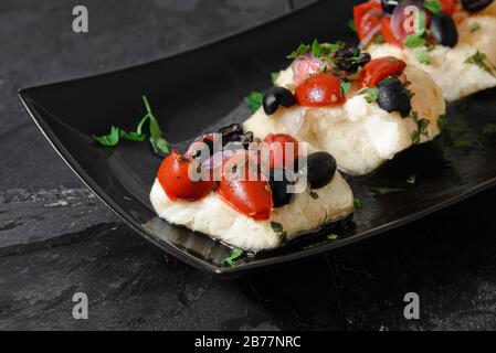 Filets de morue aux olives noires, tomates et oignons sur une plaque en céramique noire - photo studio horizontale Banque D'Images