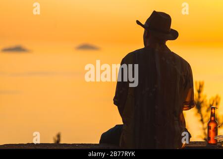 Vue sur la silhouette du dos d'un homme assis sur le mur et regardant un beau coucher de soleil avec ciel clair en arrière-plan. Banque D'Images