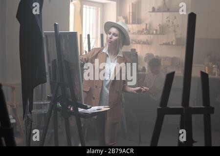 Jeune femme sérieuse en chapeau et vêtements de travail debout devant un chevalet en studio et finissant par une nouvelle peinture Banque D'Images