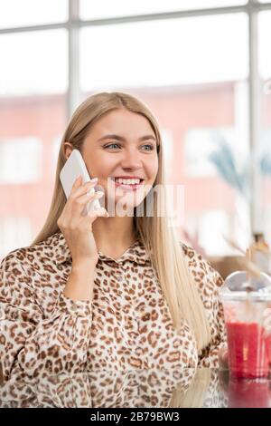 Heureuse jeune femme souriante dans un chemisier à imprimé léopard parlant sur smartphone tout en se reposant dans un café et en smoothie Banque D'Images