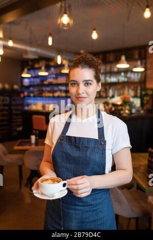 Jolie serveuse de brunette avec une tasse de cappuccino devant l'appareil photo sur fond de tables et de chaises dans le café Banque D'Images