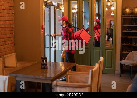 Jeune coutier avec grand sac rouge sur l'épaule va ouvrir la porte du café tout en laissant livrer des commandes aux clients Banque D'Images