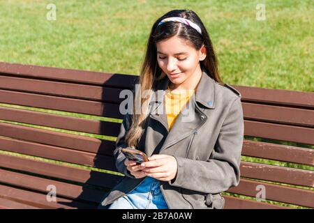 jeune belle femme hipster souriant et textant par smartphone assis sur un banc dans le parc au printemps Banque D'Images