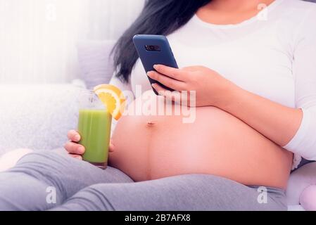 Femme enceinte utilisant son smartphone tout en se reposant sur le lit, en buvant du jus vert sain et en exposant le ventre. Banque D'Images