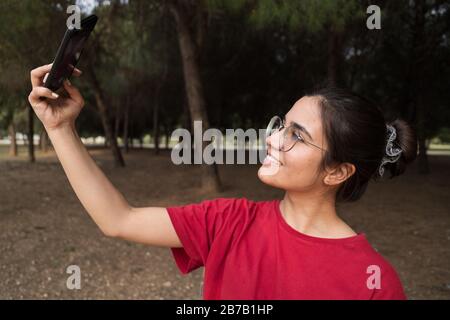 Jeune femme séduisante dans ses années vingt avec des verres et une chemise rouge assis sur un banc, tenant un téléphone mobile et prenant un selfie tout en souriant dans un Banque D'Images