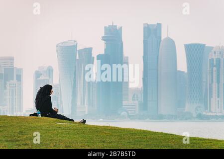 Une femme musulmane apprécie les rafraîchissements assis sur une colline herbeuse à Doha, au Qatar. La ligne d'horizon du quartier financier peut être vue à travers la brume en arrière-plan Banque D'Images