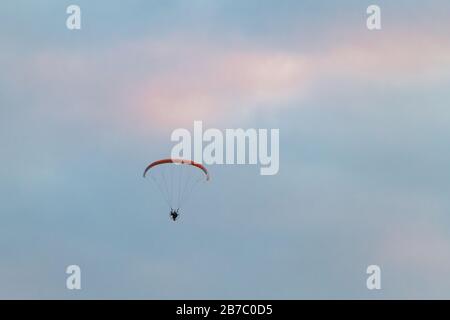 Parapente motorisé, homme volant haut dans un ciel avec parachute et moteur sur son dos. Le ciel rose sur un fond, coucher de soleil. Banque D'Images