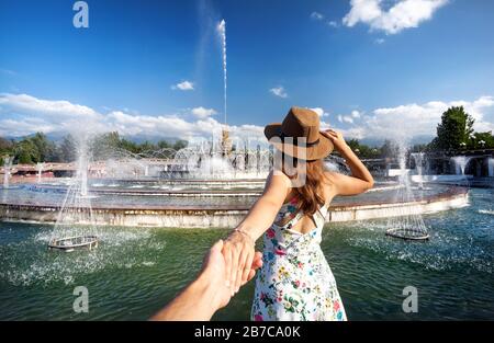Femme en robe blanche et chapeau tenant l'homme à la main et allant à la fontaine dans le parc à Almaty, Kazakhstan Banque D'Images