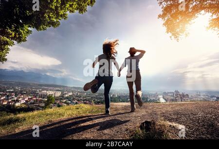Deux amis tenant les mains et courir à la vue magnifique du panorama de la ville dans les montagnes au coucher du soleil dramatique pluvieux Banque D'Images