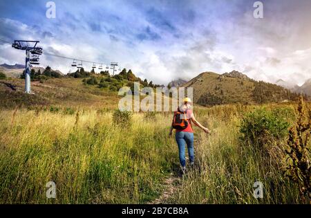 Femme de tourisme avec sac à dos et chapeau jaune touchant l'herbe près du téléphérique dans la vallée de la montagne au coucher du soleil Banque D'Images